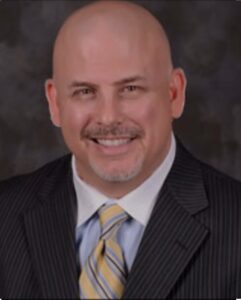 Headshot of Drew Brittain, Vice President of Sales at True Incentive, wearing a dark pinstripe suit, light blue shirt, and yellow striped tie, smiling.